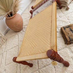 May include: A charpai daybed featuring a rectangular wooden frame with dark brown carved legs and a woven seat made of natural tan fiber in a geometric pattern. A small tan tag is attached to one of the legs, and white rope netting is visible.