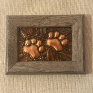 May include: A rectangular wall art piece featuring two raised copper-colored animal paw prints centered on a textured, dark metallic background. The artwork is enclosed in a simple, weathered gray wooden frame with a visible wood grain pattern.