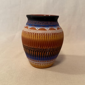 May include: A small ceramic vase featuring a black rim and a rounded body decorated with horizontal bands of blue, orange, and yellow. The surface displays intricate geometric patterns, including a Greek key design and vertical etched lines.