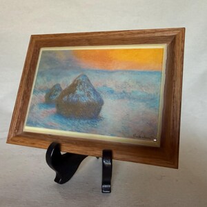 May include: A rectangular framed print featuring an impressionist landscape painting of two haystacks in a field under a golden sunset sky. The artwork is encased in a medium-toned wooden frame and is displayed on a black plastic easel stand.