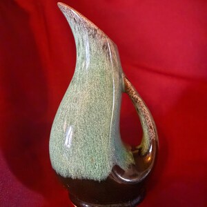 May include: A vintage ceramic pitcher with a unique, elongated shape. The pitcher features a gradient of colors, transitioning from a dark brown base to a light green body and a brown rim. The handle is curved and integrated into the design.