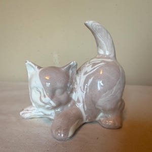 May include: A ceramic cat figurine featuring a glossy finish with a marbled pattern of soft mauve and white. The cat is depicted in a playful, crouching position with its tail held upright, showcasing smooth, rounded contours and a reflective surface.