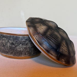 May include: A round ceramic lidded container featuring a textured gray and black exterior with a gold-toned rim. The lid is sculpted with a raised, segmented pattern resembling a shell, while the base has a mottled, stone-like finish.
