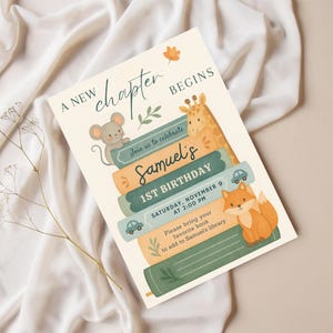 May include: An invitation card with the text "A New Chapter Begins" and "Samuel's 1st Birthday". The design features illustrations of a mouse, giraffe, and fox, with stacked books. The invitation is on a cream background.