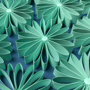 May include: A close-up of several mint green paper flowers with curved petals, arranged on a blue surface. Each flower has a central button. The flowers are evenly spaced, creating a repeating pattern.