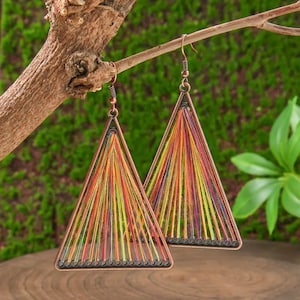 以下が含まれることがあります： カラフルな糸を使った、銅色の三角形のイヤリングのペア。枝から吊り下げられています。糸は赤、オレンジ、黄色、緑の色合いです。背景は緑色です。