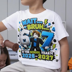 Könnte beinhalten: Ein weißes Kurzarm-T-Shirt mit einem Grafikdruck und dem Text "WAIT... BRUH... 6 7 IS THE SCHOOL YEAR?! 2026-2027". Das Design zeigt eine blockartige Spielfigur, die große Zahlen hält, umgeben von blauen und grünen Farbspritzern.