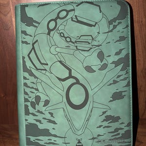 May include: Green binder featuring a detailed illustration of a dragon-like creature with a large circular design. The artwork is in a darker shade of green, set against a lighter green background. The binder has a stitched border and a small logo at the bottom.