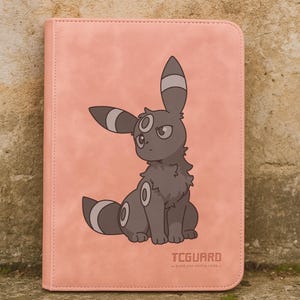 May include: Pink card binder featuring a cartoon Umbreon illustration. The Umbreon is gray with white and black accents. The text "TCGUARD - guide your trading cards -" is printed on the bottom right corner.