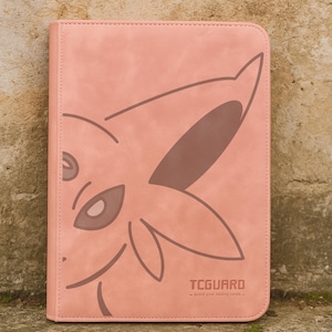 Puede incluir: Un archivador de tarjetas rosa con un diseño de personaje de dibujos animados estilizado. El archivador tiene un borde cosido y el texto "TCGUARD - guide your trading cards". El diseño es de color marrón claro.