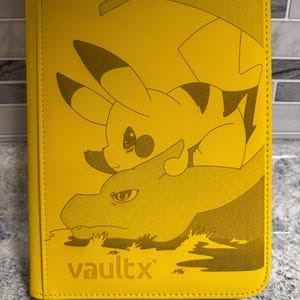 Puede incluir: Carpeta amarilla Vault X para tarjetas coleccionables con un diseño de Pikachu y Charizard. La carpeta tiene un borde cosido y cierre de cremallera. El logotipo de Vault X es visible en la parte delantera.