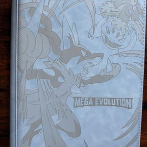 Könnte beinhalten: Ein hellblaues Sammelkartenalbum mit einem grauen, geprägten Design mit verschiedenen Mega Evolution-Charakteren. Die Worte "MEGA EVOLUTION" sind auf der Vorderseite aufgedruckt. Das Album ist mit "TCGARD" beschriftet.