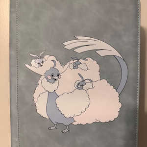 May include: A light blue rectangular binder featuring a printed graphic of a large, bird-like creature with fluffy white plumage and three smaller bird-like creatures nestled within its feathers, all set against a smooth, solid light blue background.
