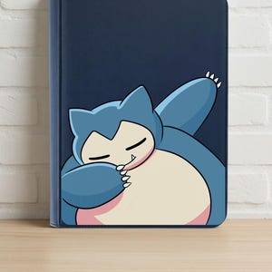 Pode incluir: Uma pasta portfólio azul-escura com uma ilustração gráfica de um personagem Snorlax em tons de azul e creme. A criatura é retratada com os olhos fechados, um braço levantado e um pequeno dente visível, sobre um fundo azul-escuro sólido.