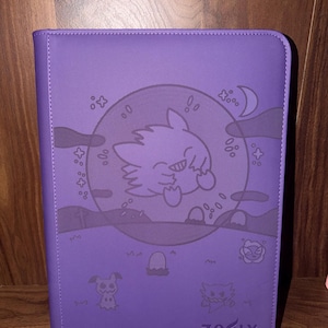 May include: A purple card binder with a cartoon graphic of a creature inside a circle, with stars, a moon, and other creatures. The word "ZOFY" is printed at the bottom.