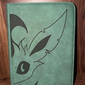 May include: A teal-colored binder with a stylized black illustration of a creature's face on the front. The design features large ears, an eye, and facial details. The text "TCGUARD" is at the bottom.