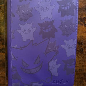 May include: Purple card binder featuring a repeating pattern of cartoon ghost characters in various shades of purple. The bottom right corner has the text "ZOF LY". The binder is made of a textured material and has a stitched border.