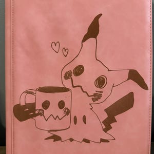 Puede incluir: Carpeta rosa para tarjetas coleccionables con una ilustración marrón de un personaje de dibujos animados con forma de fantasma y una taza de café. Dos corazones flotan sobre el personaje. El texto "TCGUARD - guard your trading cards -" está en la parte inferior.