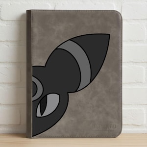 Puede incluir: Una funda gris para tableta con un diseño de gato de dibujos animados en negro y gris. El ojo y la oreja del gato son visibles en la parte delantera de la funda. La funda es rectangular y tiene una superficie texturizada. La marca está impresa en la parte inferior.