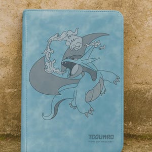 Puede incluir: Un archivador de tarjetas azul claro con un borde cosido y una ilustración gris de una criatura con forma de dragón con llamas. El texto "TCGUARD" está impreso en la parte inferior.