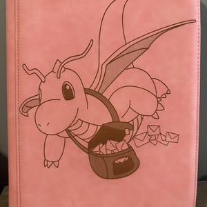 May include: Pink trading card binder with a cartoon dragon delivering mail. The binder has a stitched edge and the text "TCGUARD - guard your trading cards-" at the bottom.