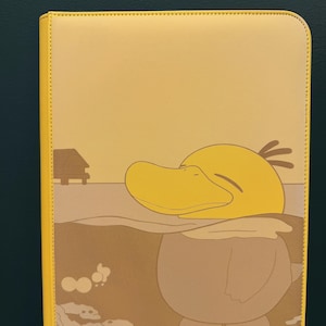 Puede incluir: Carpeta amarilla con cremallera con un diseño de pato de dibujos animados. El pato está representado en el agua con un pequeño edificio en el fondo. La carpeta tiene un borde amarillo y una superficie texturizada.
