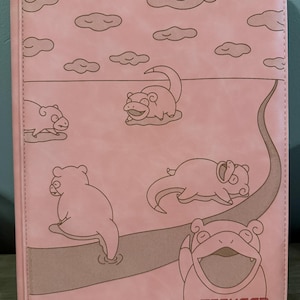 May include: Pink trading card binder featuring a cartoon design of a Slowpoke in a landscape with clouds. The binder has a brown path and the text "TCGUARD - guard your trading cards".