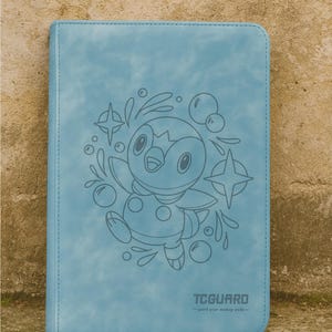 May include: A light blue, rectangular notebook with a stitched border. The cover features a cartoon illustration of a penguin with bubbles and stars. The text "TCGUARD" is printed at the bottom, with the tagline "guard your making safe".