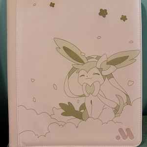 May include: A light pink binder featuring a cartoon character design. The design includes a smiling creature with large ears, sitting on a cloud. The binder has a stitched edge and a logo in the bottom right corner.