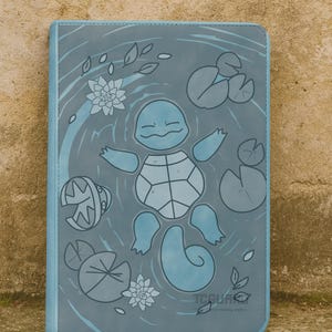 May include: A blue notebook featuring a cartoon turtle floating in water with lily pads and flowers. The notebook has a light blue border and the text "TCGUA" at the bottom.