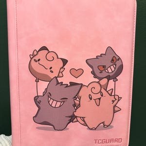 May include: Pink trading card binder featuring cartoon characters holding balloons and a heart. The binder has the text "TCGUARD - guard your trading cards" at the bottom. The binder is made of a smooth, leather-like material.