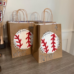 Op de afbeelding: Bruine papieren cadeauzakken met honkbalontwerpen. Elke tas heeft een witte honkbal met rode stiksels en een naam of nummer. De tassen hebben bruine gedraaide papieren handvatten en staan op een houten oppervlak.