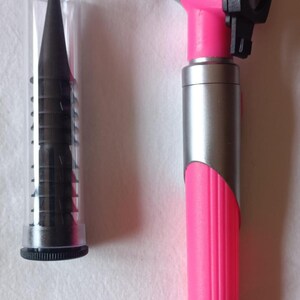 May include: A pink otoscope with a silver and black accent, and a clear container filled with black ear specula. The otoscope has a black eyepiece and a ribbed handle. Medical equipment.