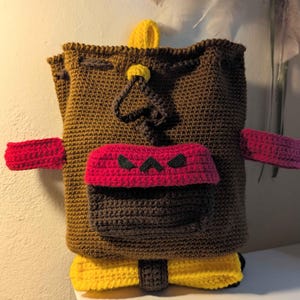 May include: A handmade crochet backpack in brown, red, and yellow. The backpack features a front pocket with a red flap and black accents, red straps, and yellow accents at the bottom. The bag has a drawstring closure.