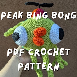 Könnte beinhalten: Eine gehäkelte grüne Kreatur mit großen weißen und schwarzen Augen, orangefarbenen Flecken und grünen Gliedmaßen. Der Text "PEAK BING BONG PDF CROCHET PATTERN" wird auf dem Bild angezeigt. Die Kreatur hat einen Propeller oben.