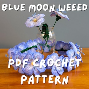 Könnte beinhalten: Eine klare Glasvase hält drei gehäkelte blaue Blumen mit gelben Zentren und grünen Stielen. Ein gehäkelter blauer Blumenkranz umgibt die Vase. Der Text "BLUE MOON WEEED" und "PDF CROCHET PATTERN" sind zu sehen.