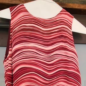 May include: Sleeveless top with a wavy striped pattern in shades of red, pink, and white. The fabric appears to be lightweight, with a flowing design. The top is hanging on a white hanger.