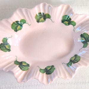 May include: A light pink, ruffled-edge ceramic dish with a central oval depression. The dish is decorated with green and yellow painted gourd-like motifs around the rim. The dish is likely vintage.