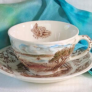 May include: Cream-colored teacup and saucer set with a landscape design in brown and blue. The cup features a handle and a detailed scene of trees, water, and a small boat. The saucer has a matching border design.