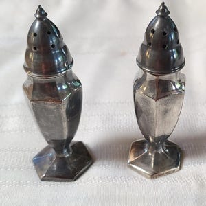 May include: A pair of antique silver salt and pepper shakers. Each shaker has a faceted body and a conical, perforated top. The shakers are displayed on a white surface. The design is classic and elegant.