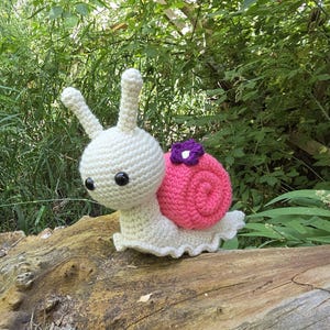 Könnte beinhalten: Eine handgefertigte Häkelschnecke mit weißem Körper, großen schwarzen Augen und einem rosa Gehäuse, das mit einer lila Blume verziert ist. Die Schnecke sitzt auf einem Holzstück, mit einem natürlichen grünen Hintergrund.