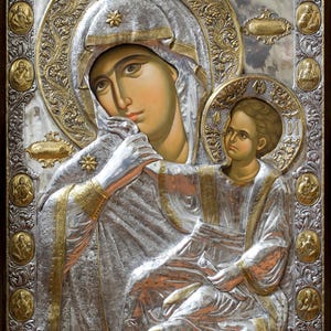 Può includere: Icona religiosa raffigurante la Vergine Maria e il Bambino. L'opera è realizzata in toni argento e oro, con intricati dettagli in rilievo e una cornice in legno scuro. Le figure sono adornate da aureole e corone.