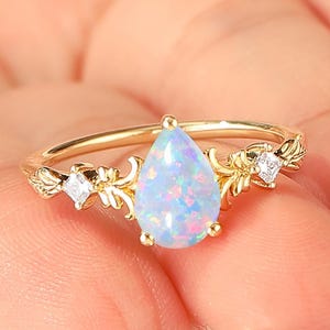 May include: A gold ring featuring a pear-shaped, light blue opal gemstone. The ring has a delicate floral design with small, clear accent stones on each side of the opal. The ring is held up by a person's fingers.