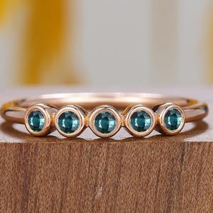 May include: A rose gold ring featuring five round, teal gemstones. The ring is set on a wooden surface with a blurred yellow and white background. The gemstones are evenly spaced along the band.