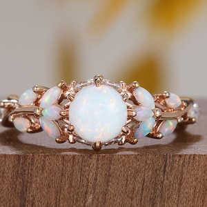 Pode incluir: Anel de ouro rosa com uma grande pedra central de opala branca redonda. O anel é adornado com opalas menores e detalhes intrincados, criando um design floral ou em forma de folha. O anel está sobre uma superfície de madeira.