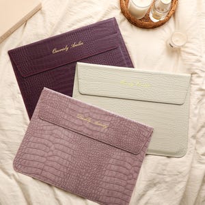 May include: Three rectangular laptop sleeves with a textured crocodile skin pattern. One sleeve is deep purple, one is light cream, and one is dusty rose. Each sleeve features a flap closure and gold-colored cursive text embroidered on the front.