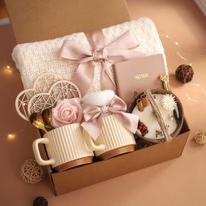K&ouml;nnte beinhalten: Eine Geschenkbox mit einer cremefarbenen Decke, einem rosa Band, einem Notizbuch, zwei Tassen, einer Kerze und dekorativen Elementen. Die Tassen sind cremefarben mit braunen B&ouml;den. Die Kerze befindet sich in einer Holzschale.