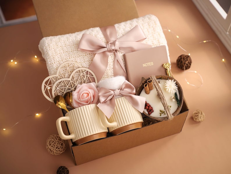 K&ouml;nnte beinhalten: Eine Geschenkbox mit einer cremefarbenen Decke, einem rosa Band, einem Notizbuch, zwei Tassen, einer Kerze und dekorativen Elementen. Die Tassen sind cremefarben mit braunen B&ouml;den. Die Kerze befindet sich in einer Holzschale.