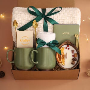 K&ouml;nnte beinhalten: Eine Geschenkbox mit einer gr&uuml;nen Tasse, einer wei&szlig;en Decke, einer Kerze, einem Notizbuch und einem Schokoriegel. Die Tasse hat vertikale Linien und einen Henkel. Die Kerze befindet sich in einer Holzschale mit getrockneten Orangenscheiben. Die Box ist mit einem gr&uuml;nen Band gebunden.
