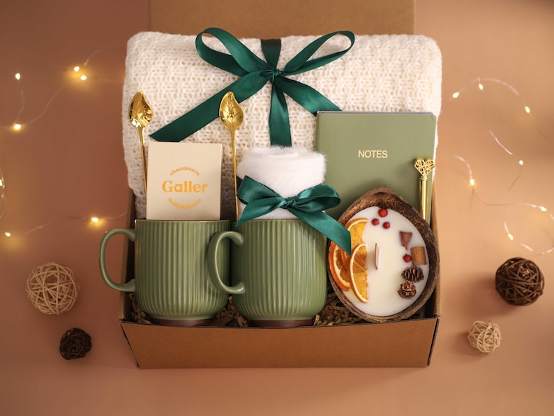 K&ouml;nnte beinhalten: Eine Geschenkbox mit einer gr&uuml;nen Tasse, einer wei&szlig;en Decke, einer Kerze, einem Notizbuch und einem Schokoriegel. Die Tasse hat vertikale Linien und einen Henkel. Die Kerze befindet sich in einer Holzschale mit getrockneten Orangenscheiben. Die Box ist mit einem gr&uuml;nen Band gebunden.